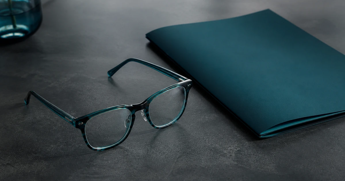 Teal and charcoal editorial still of unmarked eyeglasses and a closed folder, no people or text overlay